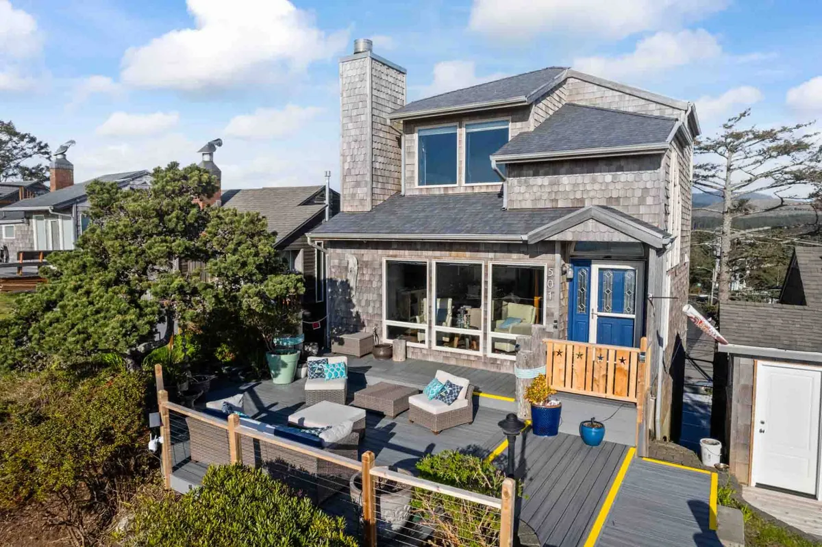 An inviting front exterior view of the property, showing a welcoming porch seating area, large windows, a blue door, and decorative plants. The setting suggests a blend of comfort, style, and a connection to the outdoors.