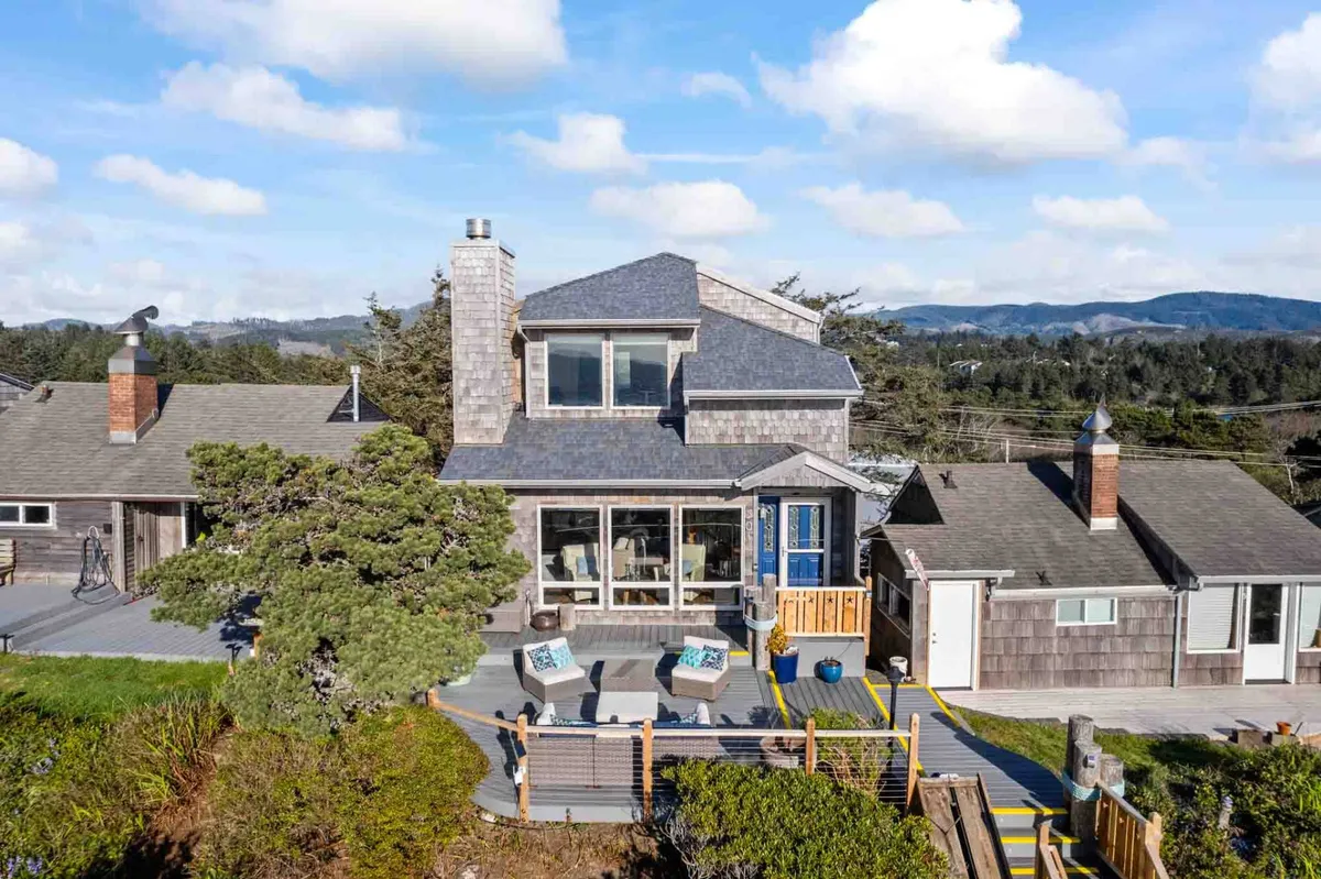 The image showcases the front of Sea Star Villa, a single-family house nestled on a lush, landscaped hilltop. The inviting front porch features comfortable seating perfect for relaxing, while expansive windows hint at spectacular ocean views inside. The classic shingle siding and vibrant garden beds create a charming, coastal ambiance.