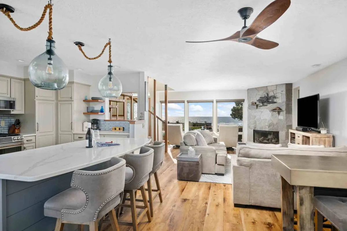 A seamless, open-concept space where the kitchen island flows into a living room seating area, each bathed in natural light from oversized windows. The fireplace and well-appointed kitchen foster both intimate gatherings and lively celebrations, with glimpses of the landscape beyond.