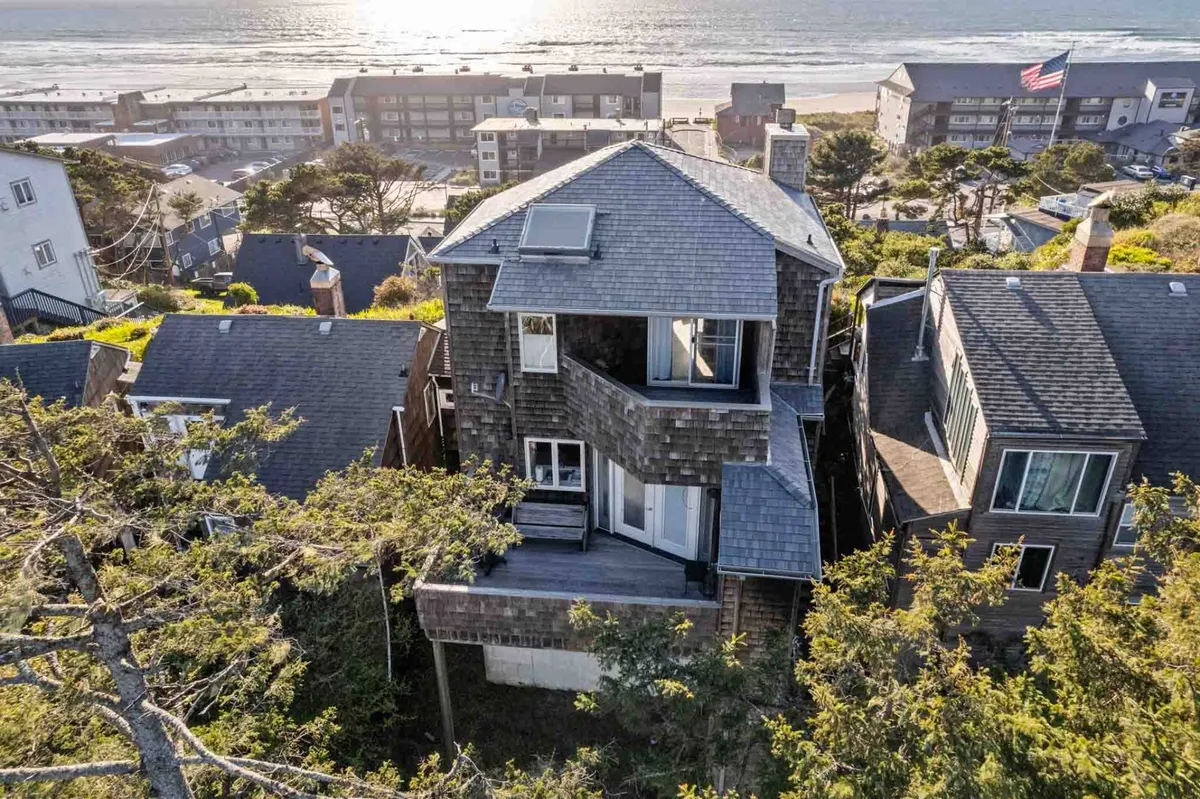 This image captures the unique architecture of the house set against a backdrop of surrounding trees and offers a clear view of the ocean. The spacious deck, ideal for relaxing and enjoying coastal views, visually connects the property with the beach just moments away.