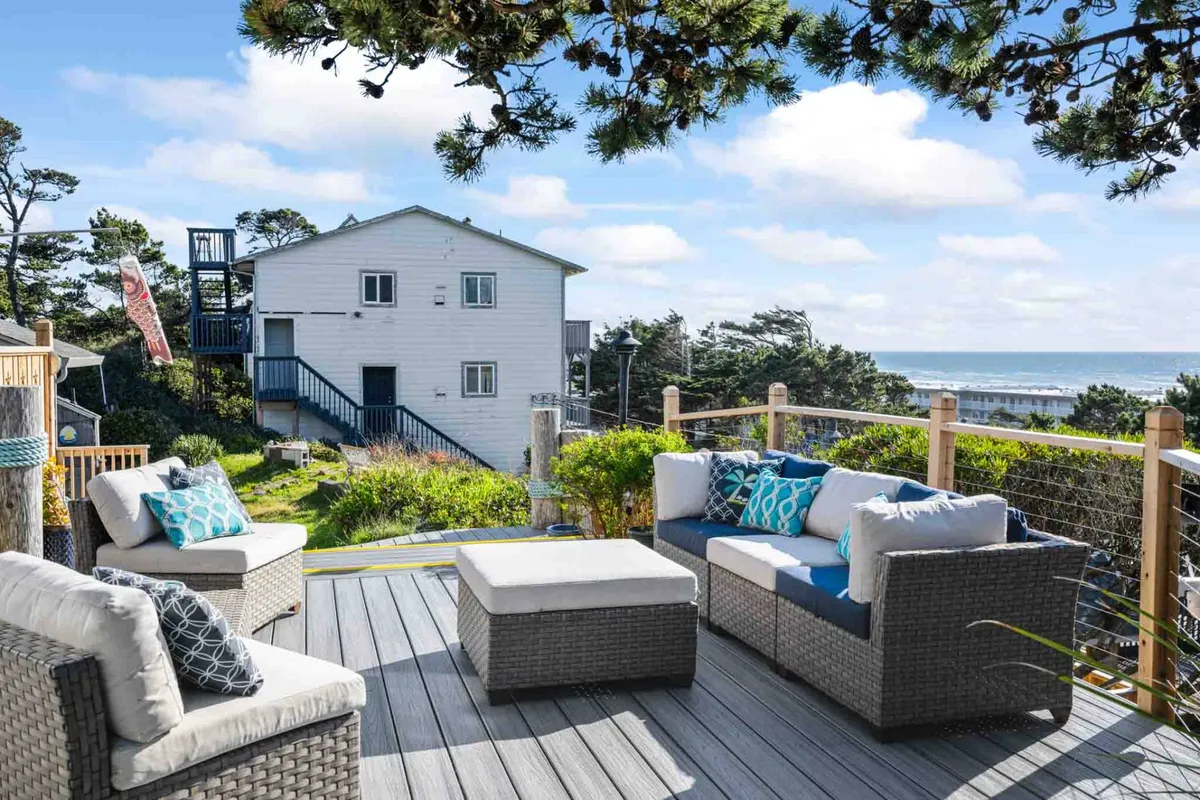 A serene deck area furnished with cozy seating overlooks the ocean, surrounded by greenery. The expansive deck is set up for both quiet moments with a book and lively gatherings with friends or family, capturing the essence of lakeside and beachside relaxation.