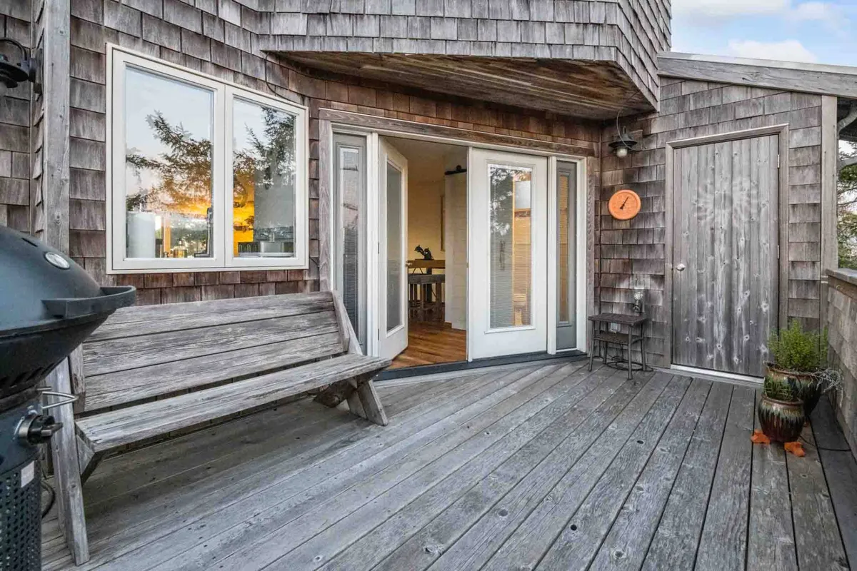 An exterior shot showing a wooden deck with a grill, bench seating, and planters. The space is set up for outdoor dining and relaxation, perfect for barbecues or morning coffee. Double doors suggest seamless access from the interior to the deck.