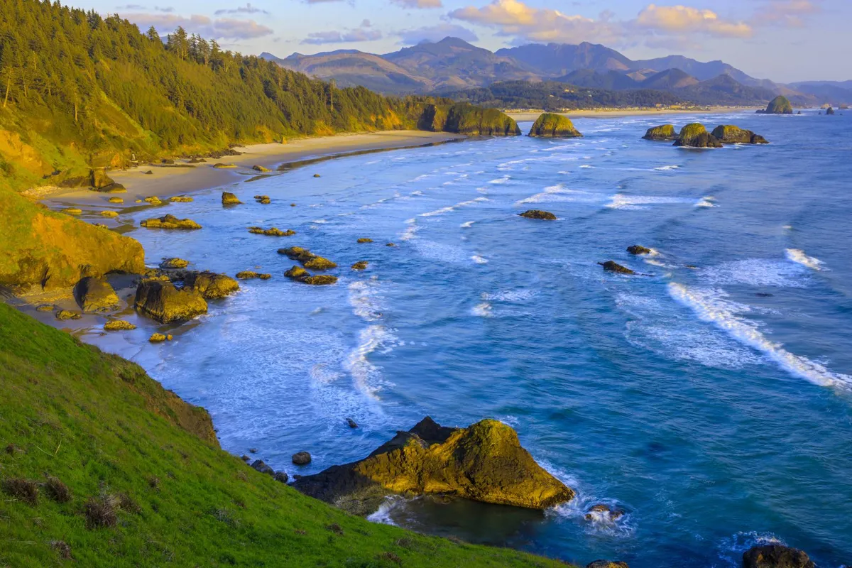 A sweeping coastal landscape featuring dramatic ocean waves, rocky formations, distant mountains, sandy beach, and coastal forest. The scene is tranquil and expansive, evoking the sense of adventure and natural beauty found along Highway 101 and the Oregon Coast.